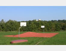 Freiluft-Basketballplatz mit rotem Asphaltbelag auf einer grünen Wiese, umgeben von Bäumen und mit klarem Himmel im Hintergrund. Zwei Basketballkörbe stehen an den gegenüberliegenden Enden des Spielfelds.