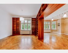 Großzügiger Wohnraum mit Holzboden in Fischgrätmuster, großen Fenstern mit Blick auf grüne Baumkronen, sowie integrierten hölzernen Bücherregalen und einer Raumtrennung aus Holz. Die Wände sind teils verputzt, und eine Hängelampe hängt zentral im Vorderraum.