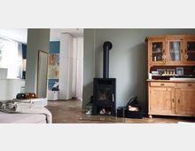 Wohnzimmer mit hellgrüner Wand, einem schwarzen Kaminofen auf einer Glasplatte und einem hölzernen Vitrinenschrank rechts. Im Hintergrund sind eine offene Küche, ein großer Spiegel und moderne Kunst zu sehen, mit Tageslicht durch weiße Vorhänge.