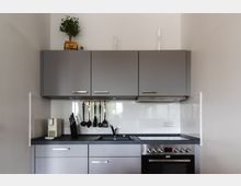 Moderne Küche mit grauen Oberschränken, einem schwarzen Arbeitsbereich und weißen Fliesen an der Wand. Unter den Schränken hängen Küchenutensilien, daneben ein eingebauter Herd und eine Spüle.