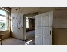 Ein verlassenes Zimmer mit einer offenen weißen Tür, die in einen weiteren leeren Raum führt. Die Wände sind abgenutzt, mit freigelegtem Putz und Kabeln, und große, rechteckige Fenster lassen Tageslicht herein, das auf den Holzboden fällt.