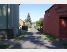 Schmale, asphaltierte Straße in einem Dorf oder Wohngebiet, gesäumt von einem grünen Gebäude links und einem roten Haus mit Ziegeldach rechts. Im Hintergrund ist eine Baumreihe sichtbar, und entlang der Straße stehen vereinzelt Gartenzäune und Laternen.