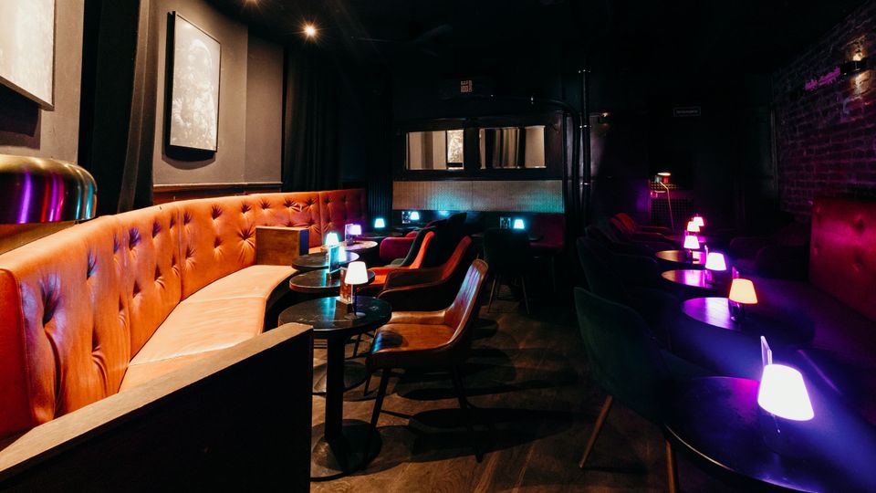 Dunkler Bar- oder Lounge-Innenraum mit einer langen, gepolsterten Lederbank, kleinen runden Tischen und Sesseln. Gedimmte Deckenbeleuchtung und mehrere farbige Tischlampen sorgen für Akzente, dazu Backsteinwand und schwere Vorhänge.