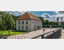 Ein zweistöckiges Gebäude mit weißer Fassade und rotem Satteldach, das als Tourist-Information und Bibliothek gekennzeichnet ist, steht an einem gepflasterten Platz mit Bänken und Grünflächen. Im Hintergrund sind Bäume, geparkte Autos und ein Kirchturm zu sehen.