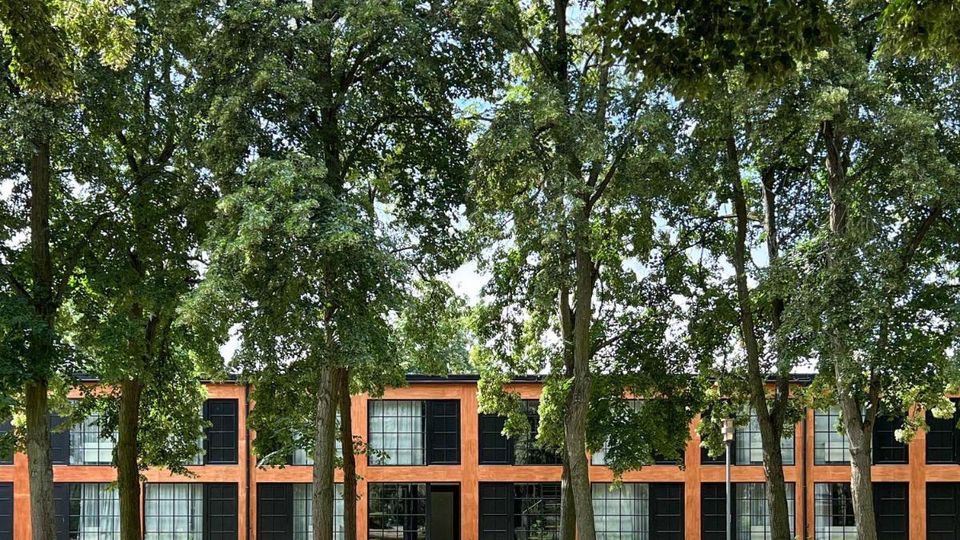 Zweistöckiges Gebäude mit einer Fassade aus orangefarbenem Backstein und großen schwarzen Fenstern, umgeben von hohen Bäumen in einem parkähnlichen Umfeld. Ein Weg führt entlang der Vorderseite des Gebäudes durch die Grünfläche.