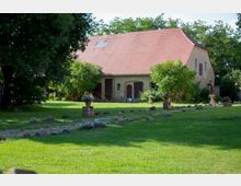 Ein ländlicher Hof mit einem großen gelben Gebäude, das ein rotes Satteldach und braune Holztüren hat. Der Vordergrund zeigt eine gepflegte Rasenfläche mit gepflastertem Weg, dekorativ eingerahmt von Steinen und Blumentöpfen.