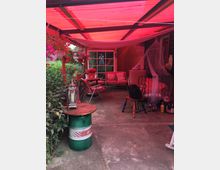 Eine überdachte Terrasse mit rotem Lichtfilter, ausgestattet mit Möbeln wie gepolsterten Stühlen, Sofa und einem improvisierten Tisch aus einem grünen Metallfass mit Holzplatte. Im Hintergrund sind Blumen, ein Fenster und verschiedene dekorative Objekte sichtbar, die den Raum gemütlich gestalten.