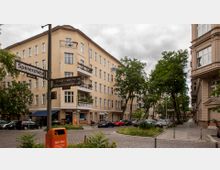 Straßenecke mit Wohnhäusern in einem städtischen Bereich, umgeben von Bäumen. Die Gebäude haben gelbe und beige Fassaden mit mehreren Balkonen, und Straßenschilder markieren die Kreuzung von Spenerstraße und Melanchthonstraße. Im Vordergrund steht ein orangefarbener Mülleimer auf einem gepflasterten Gehweg.