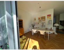 Heller Wohnraum mit einem Esstisch und vier Designerstühlen in der Mitte, einer hölzernen Vitrine an der Wand und gerahmten Kunstwerken und Postern an der weißen Wand. Ein modernes, kugelförmiges Hängeleuchten-Design hängt über dem Tisch, während links Tageslicht durch eine geöffnete Glastür auf den Fischgrät-Holzboden fällt.