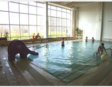 Hallenbad mit rechteckigem Schwimmbecken, umgeben von Fliesenboden. Große Fensterfront lässt Tageslicht herein, im Becken schwimmen mehrere Menschen, eine kleine lila Rutsche befindet sich am Beckenrand.