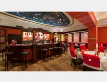 Ein festlich dekoriertes Restaurant mit roten Wänden und Holzboden, einem großen Tresen mit Barhockern und gedeckten Tischen, die mit roten Stuhlhussen verziert sind. Die Decke ist mit einem gemalten Himmelmotiv versehen, und die Bar ist mit Glasregalen und beleuchteten Flaschen ausgestattet.