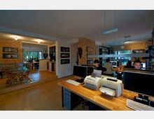 Eine moderne Büroeinrichtung mit mehreren Arbeitstischen und Computern, umgeben von Regalen mit Büromaterialien und einer Ziegelwand im Hintergrund. Im Vordergrund ein Schreibtisch mit Druckern und Monitoren, im hinteren Bereich Personen, die an Rechnern arbeiten, sowie ein angrenzender Besprechungsbereich mit einem Glastisch und Holzstühlen.