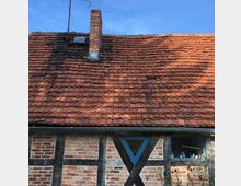 Fachwerkhaus mit einer Fassade aus roten Backsteinen und dunklen Holzbalken, darüber ein geneigtes Dach mit roten Ziegeln und einem gemauerten Schornstein. Im unteren Bereich ist ein blau gerahmtes Fenster sowie ein kleines dreieckiges Oberlicht sichtbar.