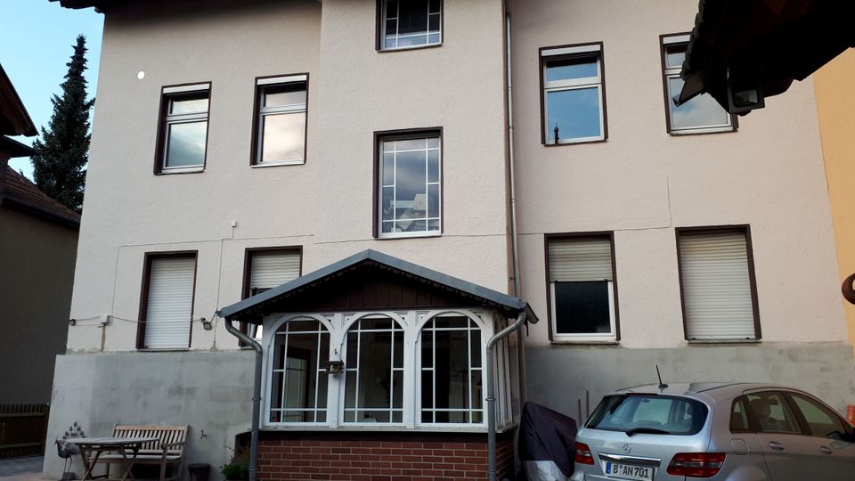 Hinterhofansicht eines mehrstöckigen Wohnhauses mit heller Putzfassade, mehreren rechteckigen Fenstern und einem kleinen verglasten Anbau mit Satteldach und Ziegelsockel. Im gepflasterten Hof stehen ein geparktes Auto, daneben eine Bank mit Tisch sowie einige Pflanzkübel.