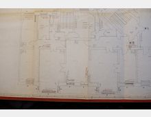 Grundrisszeichnung eines Gebäudes mit zahlreichen Räumen, Treppen und Türen, beschriftet mit Maßangaben und handschriftlichen Notizen in rot und schwarz. Die Zeichnung zeigt detaillierte Raumaufteilungen, darunter Küchen- und Sanitärbereiche, sowie Maßlinien entlang der Wände.