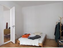 Ein schlichtes Schlafzimmer mit Holzboden und weißen Wänden. Im Raum stehen ein einfaches Bett mit Bettwäsche und Kleidung darauf, ein geöffneter Türdurchgang zu einem weiteren Zimmer sowie eine Holzgarderobe mit hängender Kleidung.