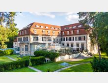 Große historische Villa mit cremefarbener Fassade und rotem Ziegeldach, umgeben von gepflegtem Garten. Auffällig ist ein großer Glasanbau mit Terrasse und Sitzmöglichkeiten, flankiert von hohen Bäumen.