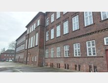 Backsteinfassade eines mehrstöckigen Gebäudes mit zahlreichen rechteckigen weißen Fensterrahmen. Im Vordergrund befindet sich eine asphaltierte Fläche, im Hintergrund sind weitere Gebäude und einige geparkte Autos zu sehen.