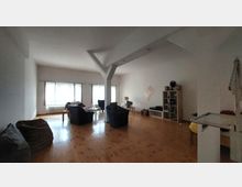 Wohnzimmer mit hellen Wänden, Holzboden und großen Fenstern, die natürliches Licht hereinlassen. Der Raum ist mit mehreren Sesseln, einem Sofa, einem Regal an der Wand und einem kleinen Tisch möbliert.