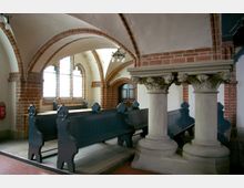 Innenraum einer Kirche mit spitzbogigen Backsteinbögen, verzierten Säulen und hölzernen Kirchenbänken. Die Wände sind weiß verputzt, und es gibt mehrere Fenster mit Bogenform, die natürliches Licht einlassen.