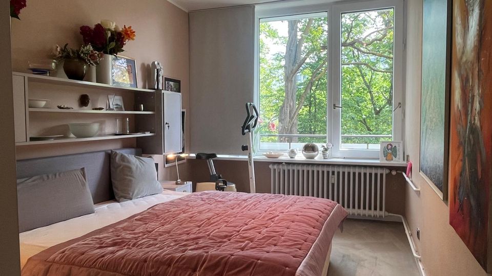 Modernes Schlafzimmer mit Doppelbett und rosafarbener Tagesdecke, wandmontierten Regalen und großformatigen Bildern an der Wand. Ein großes Fenster mit Heizkörper darunter bietet Blick auf grüne Bäume und lässt viel Tageslicht in den Raum.