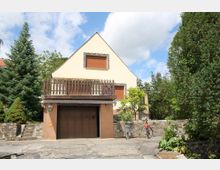 Einfamilienhaus mit Satteldach, cremefarbener Fassade und Holzbalkon über einer Garage mit braunem Tor. Umgeben von Bäumen, Sträuchern und einer niedrigen Steinmauer; zwei Fahrräder stehen vor dem Haus.