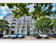 Mehrstöckiges Wohngebäude mit moderner Fassade und großen, abgerundeten Fenstern an den seitlichen Erkern. Davor geparkte Autos und Gehweg; im Vordergrund rahmen Bäume die Szene ein.