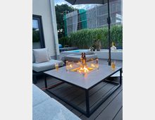 Moderne Terrasse mit grauer Holzoptik, einer Feuerstelle in einem rechteckigen Tisch mit Glasumrandung und Sitzgelegenheiten mit hellgrauen Polstern. Im Hintergrund sind hohe Ziergräser, ein Pool sowie ein mit Baugerüsten verkleidetes Gebäude zu sehen.