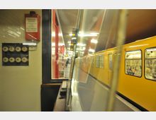 U-Bahnhof mit gelber U-Bahn an einem Bahnsteig, reflektiert in einer Glasscheibe; links ein rotes Notruf-Schild und alte Stromanschlüsse an der Wand.