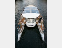 Ein Katamaran mit Solarpanelen auf dem Dach fährt auf einem Gewässer, umgeben von mehreren Personen, die auf den seitlichen Decks stehen. Der Schriftzug "Solon" ist an der Front des Bootes zu sehen.