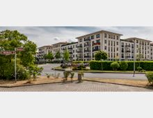 Moderne Wohnanlage mit mehreren mehrstöckigen Gebäuden in heller Fassade, Balkonen und roten Dächern, umgeben von gepflegten Grünflächen und einer Straße im Vordergrund. Links im Bild befindet sich ein Straßenschild mit den Namen "Wildbahn" und "Blumenkorso".