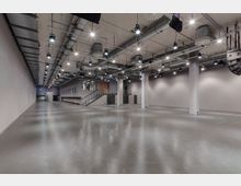 Großer, moderner Innenraum mit Betonboden, blanken Wänden und einer offenen Decke mit sichtbaren Lüftungsrohren und Beleuchtungsanlagen. Ein erhöhtes Podest mit einer Treppe, mehreren Türen und Glasfenstern befindet sich an einer Wand.
