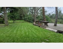 Grüne Rasenfläche mit einem erhöhten Holzdeck, auf dem ein Tisch und eine Bank stehen, umgeben von Bäumen und Sträuchern in einer Gartenlandschaft. Im Hintergrund sind weitere Bäume und ein kleines Gebäude mit heller Fassade zu sehen.