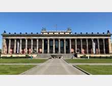 Frontale Ansicht des Alten Museums in Berlin, einem neoklassizistischen Gebäude mit einer Fassade aus korinthischen Säulen, einer zentralen Treppe und Skulpturen auf beiden Seiten. Im Vordergrund befindet sich eine Rasenfläche mit einem gepflasterten Weg.