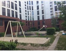 Innenhof eines modernen Wohngebäudes mit Spielplatz. Zu sehen sind eine Schaukel, ein kleiner Sandkasten, eine Rutsche sowie Bänke, umgeben von Grünflächen und Gebäudefassaden aus weißem Putz und rotem Backstein.