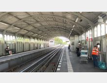 Überdachter S-Bahnhof mit gewölbtem Metalldach und seitlichen Fensterreihen, zwei Gleisen in der Mitte, Sitzbänken auf beiden Bahnsteigen und vereinzelten Personen, darunter ein Mitarbeiter in einer orangefarbenen Warnweste. Im Hintergrund sind Lichtanzeigen und Bäume sichtbar.