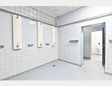 Hell gefliester Duschraum mit weißen, wandmontierten Duscheinheiten und kleinen, quadratischen Fliesen auf Boden und Wänden; im Hintergrund ist ein angrenzender Waschraum mit Urinalen sichtbar.