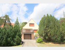 Ein Einfamilienhaus mit Satteldach und hellem Putz, umgeben von dichten Bäumen und Sträuchern. Vor dem Haus befindet sich ein gepflasterter Weg, der zu einer Garage im Untergeschoss führt, während die obere Etage mit einem Balkon ausgestattet ist.