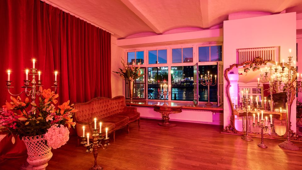 Eleganter Innenraum mit roten Samtvorhängen, antikem Sofa, poliertem Holzboden und zahlreichen Kerzenleuchtern sowie Blumenarrangements. Große Fensterfront mit Blick auf ein beleuchtetes Ufer bei Nacht und ein großer vergoldeter Spiegel an der Wand.