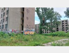Verlassene Wohnblöcke aus Beton mit beschädigten Fassaden und teils zerstörten Fenstern, umgeben von wilder Vegetation und Graffiti an den Wänden. Im Vordergrund befindet sich eine mit Gras und Wildblumen bewachsene Fläche, daneben ein verwitterter Gehweg.