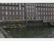 Backsteingebäude in symmetrischer Architektur mit weißen Fenstern und einem Steineingang, gelegen direkt an einer Wasserstraße mit gemauerter Uferbefestigung und einer Brücke rechts im Bild.