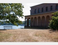 Ein Gebäude mit rot-weißer Ziegelarchitektur und Rundbogenarkaden steht am Ufer eines Sees. Im Vordergrund sind eine trockene Wiese, ein Baum und eine Holzbank zu sehen, während im Hintergrund ein Boot auf dem Wasser fährt.