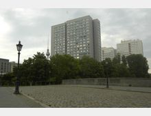 Mehrstöckige Wohngebäude vor einer Baumreihe und gepflastertem Platz, mit historischen Straßenlaternen im Vordergrund. Im Hintergrund ragt der Berliner Fernsehturm in den bewölkten Himmel.