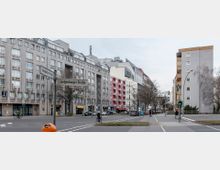 Straßenszene an der Kreuzung von Lietzenburger Straße und Ansbacher Straße in einer Stadt, mit Mehrfamilienhäusern im Hintergrund, darunter graue und farbige Fassaden. Im Vordergrund ein Fußgängerüberweg, Straßenlaternen, und ein Verkehrsschild mit den Straßennamen.