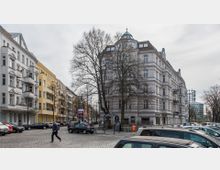 Straßenszene mit historischen Wohngebäuden im Berliner Altbaustil, darunter ein Eckhaus mit hellgrauer Fassade und einem Biergarten im Erdgeschoss. Im Vordergrund sind parkende Autos und eine gepflasterte Straße zu sehen, daneben karge Bäume und ein Passant.