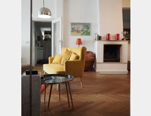 Wohnzimmer mit einem Holzboden in Fischgrätenmuster, einem gelben Sessel mit gemustertem Kissen, einem kleinen runden Beistelltisch und einem Kamin an der weißen Wand. Im Hintergrund befindet sich eine Tür, die zu einem Flur mit Treppe führt.