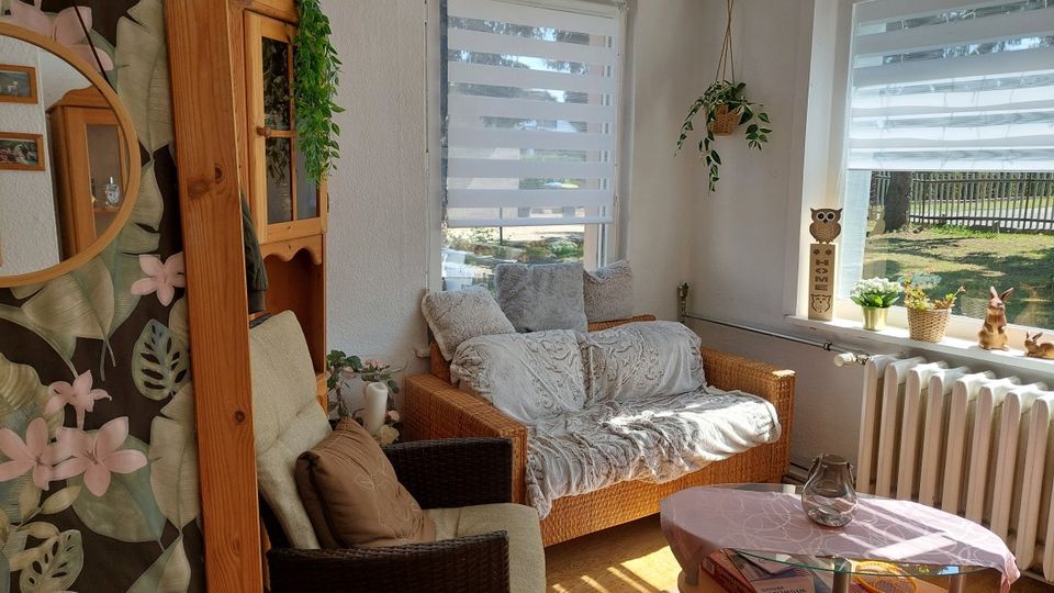 Heller Wohnbereich mit Rattansofa und Sessel, dekoriert mit Kissen, Decken und Pflanzen, neben zwei großen Fenstern mit Doppelrollos. Vor den Fenstern steht ein Heizkörper; auf der Fensterbank befinden sich kleine Dekofiguren und Topfpflanzen, davor ein kleiner Couchtisch.