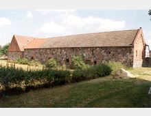 Historisches landwirtschaftliches Gebäude mit Wänden aus grobem Feldsteinmauerwerk und einem Satteldach mit Ziegeldeckung. Im Vordergrund sind Rasenflächen und Büsche zu sehen; der Himmel ist teils bewölkt.