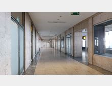 Langer, heller Flur in einem öffentlichen Gebäude mit beige gefliesten Böden und Wänden aus hellem Stein. Links und rechts befinden sich Fensterfronten und Türen, darüber Notausgangsschilder und vereinzelte Feuerlöscher.