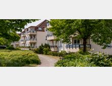 Mehrfamilienhäuser mit hellen Fassaden und roten Dachziegeln, umgeben von viel Grün und einem gepflasterten Gehweg. Vor den Balkonen befinden sich Büsche und Bäume, die den Wohnbereich abschirmen.